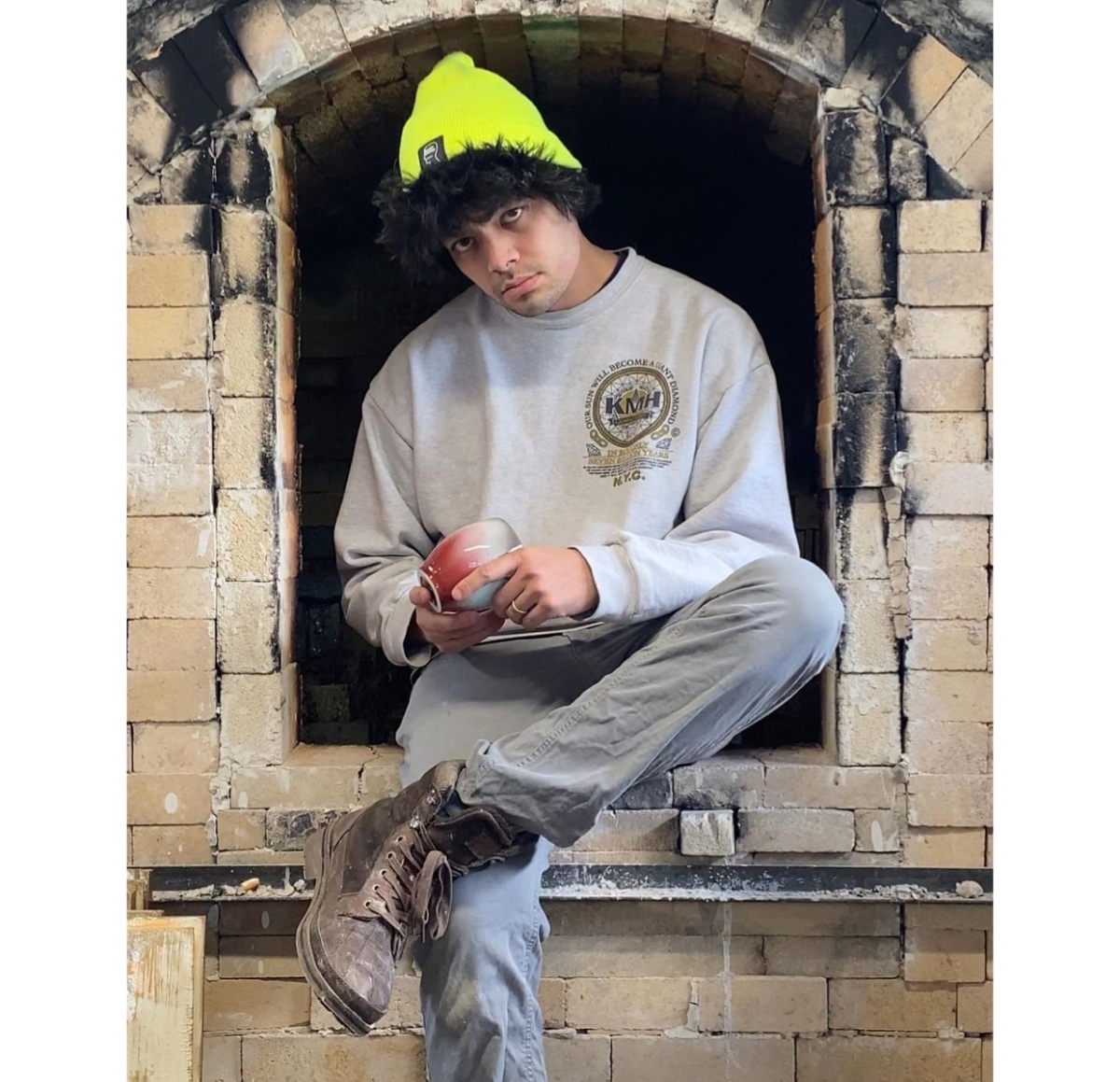 Justin Paik Reese seated in a kiln opening, holding a small glazed pot